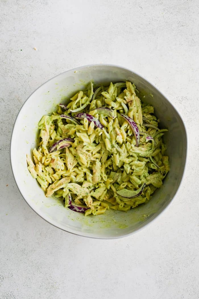 Large white mixing bowl filled with mixed together cooked pasta, shredded chicken, and sliced red onion with green goddess dressing.