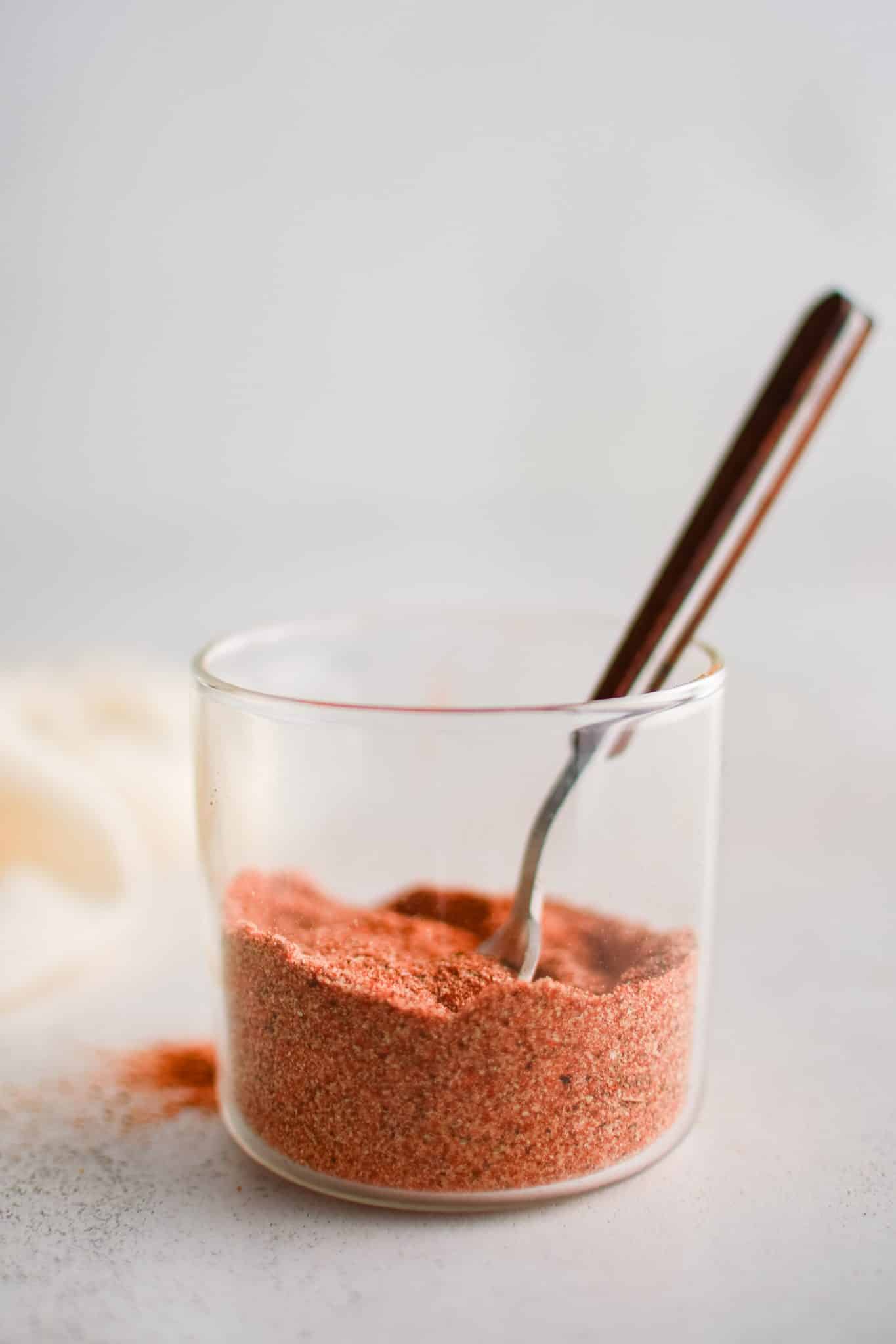 Spoon set inside a small clear glass cup filled with mixed together homemade Cajun spice blend. 