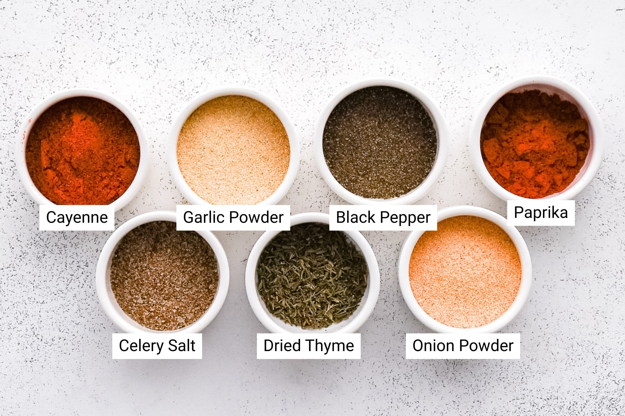 Eight small white ramekins filled with homemade Cajun seasoning ingredients arranged on a light textured background. Each ramekin is labeled with the following spices: cayenne pepper, garlic powder, black pepper, paprika, celery salt, dried thyme, and onion powder.