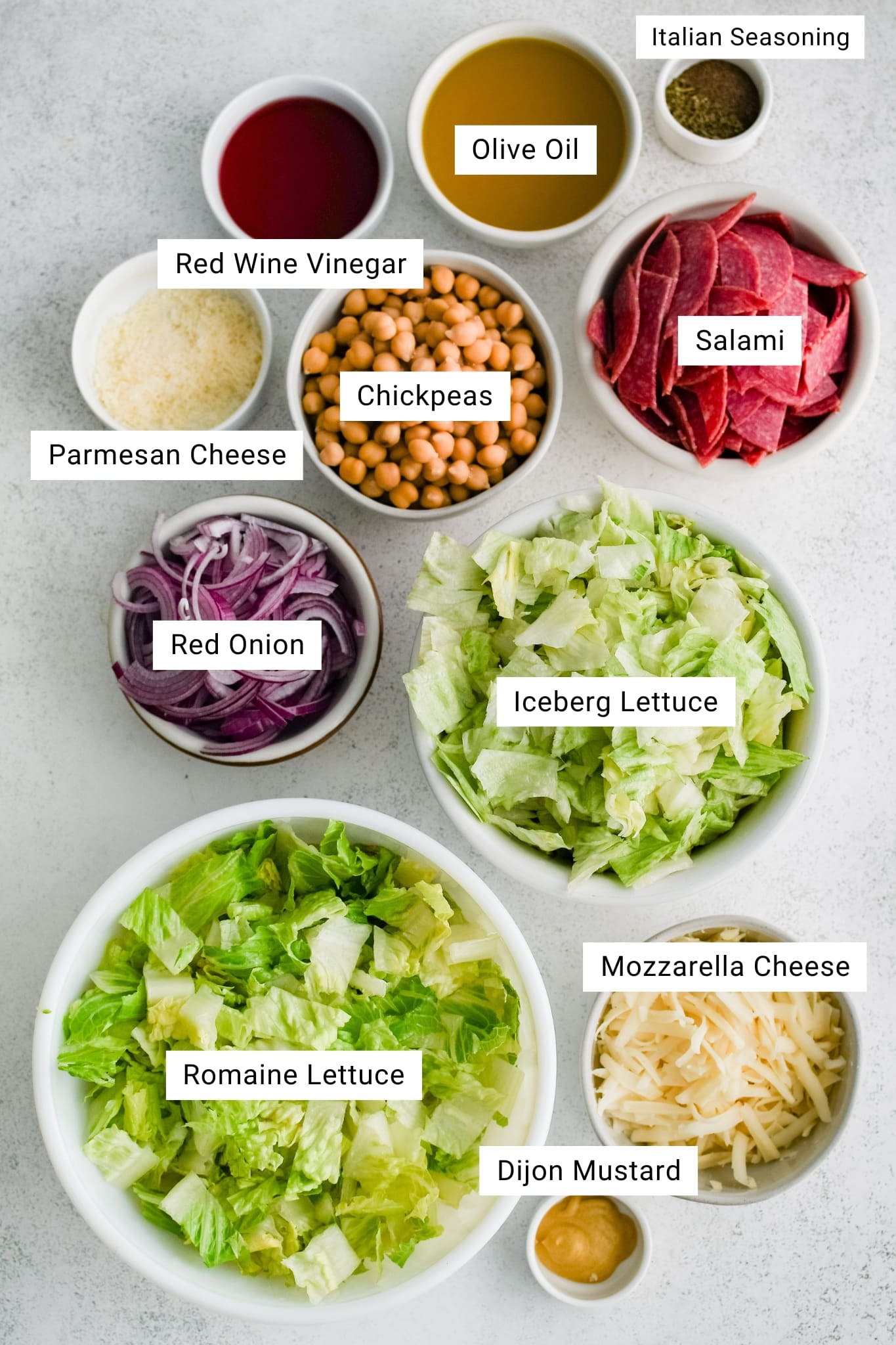 Overhead image showing labeled ingredients in small bowls for La Scala chopped salad, including olive oil, red wine vinegar, Italian seasoning, salami, Parmesan cheese, chickpeas, red onion, iceberg lettuce, romaine lettuce, mozzarella cheese, and Dijon mustard.
