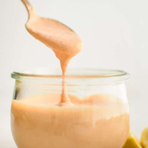 Small spoon filled with creamy dripping remoulade sauce hovering above a small round glass filled with the remaining Cajun-style remoulade sauce.