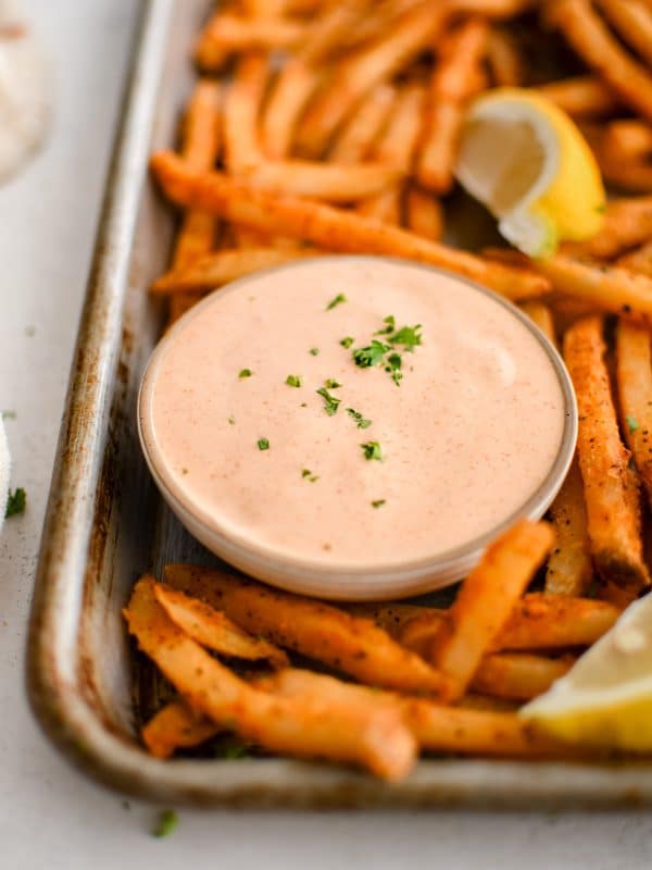 Baking sheet filled with crispy sweet potato fries, lemon wedges, and a small glass bowl filled with spicy, creamy, tangy remoulade sauce.