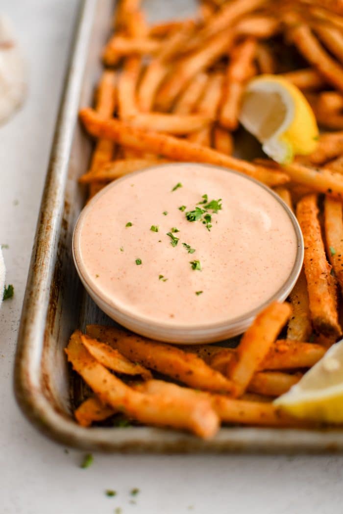 Baking sheet filled with crispy sweet potato fries, lemon wedges, and a small glass bowl filled with spicy, creamy, tangy remoulade sauce.