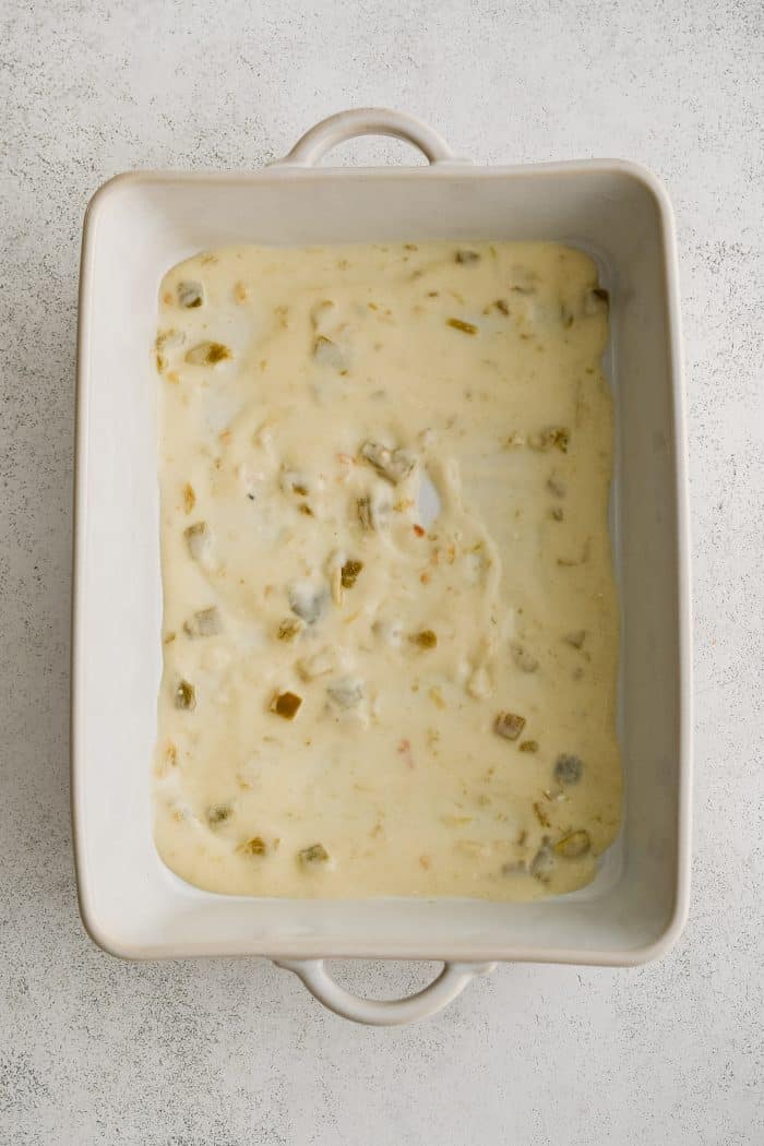 A large white baking dish spread with a thin layer of cheesy sour cream and green chili sauce.