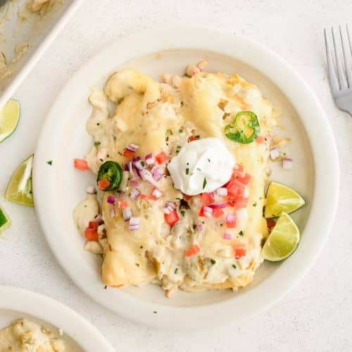 White serving plate with two sour cream chicken enchiladas topped with cheese, diced tomatoes, diced red onion, and sliced jalapeno.