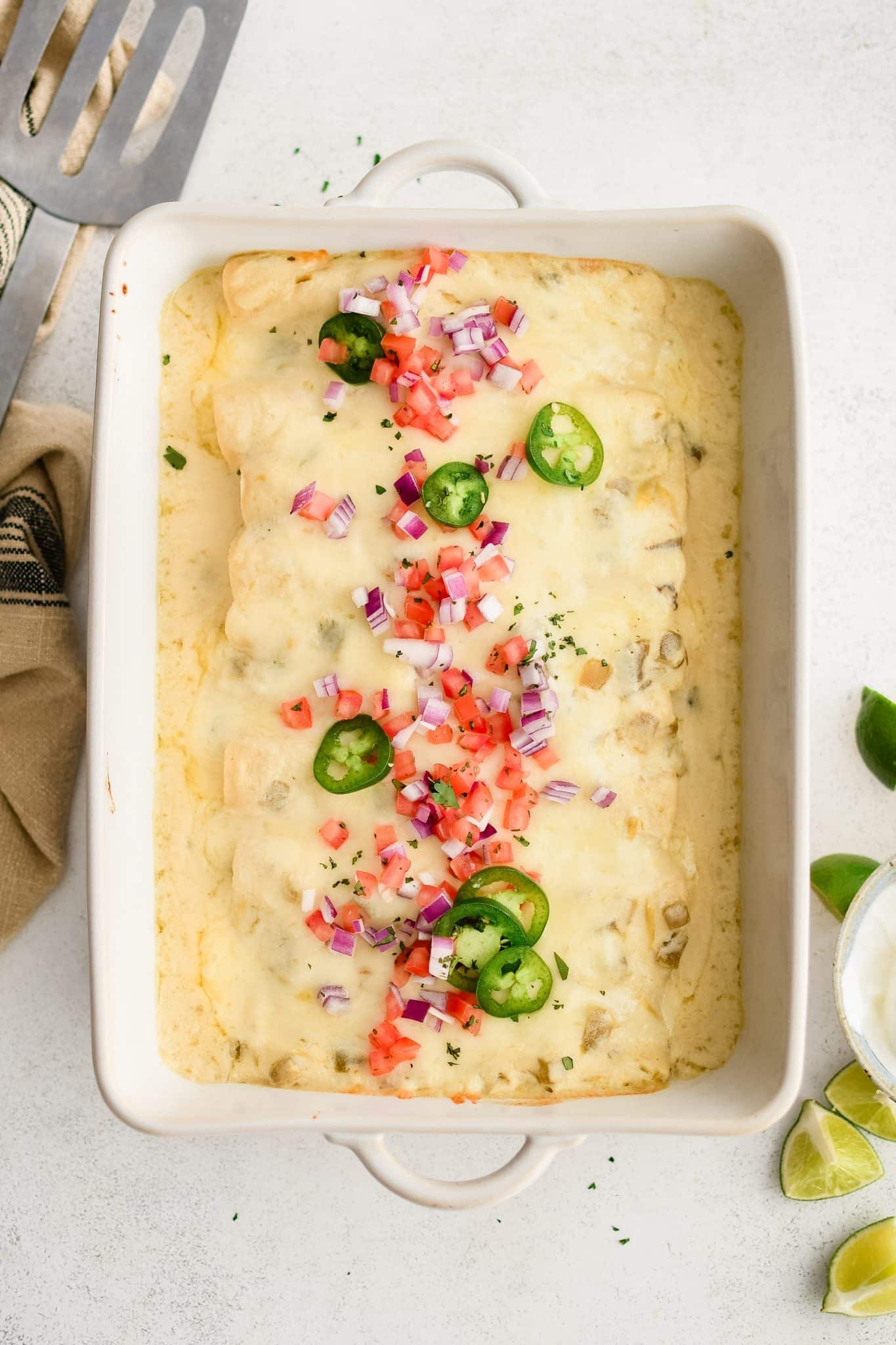 Large white casserole baking dish filled with sour cream enchiladas smothered in a creamy sour cream and cheese sauce and topped with diced red onion, diced tomatoes, and sliced jalapenos.