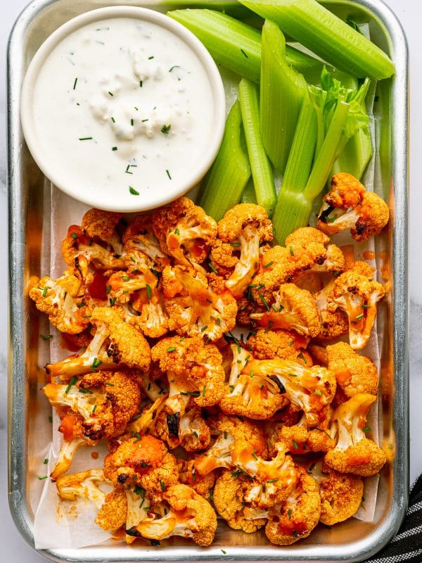 Overhead serving photo of baked buffalo cauliflower in a small metal tray lined with parchment paper, topped with chopped green chives.