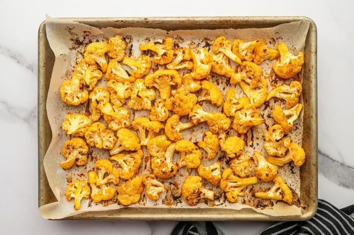 Overhead photo of baked buffalo cauliflower florets on a parchment-lined metal baking sheet with lightly browned and charred edges, showing baked-on sauce spots on the parchment paper.