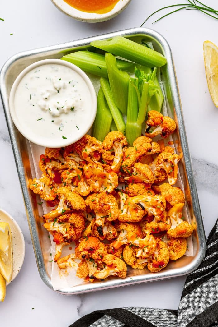 Overhead serving photo of baked buffalo cauliflower in a small metal tray lined with parchment paper, topped with chopped green chives, served beside celery sticks and a white ramekin of creamy ranch dressing, with a lemon wedge on the right and a bowl of buffalo sauce partially visible at the top.