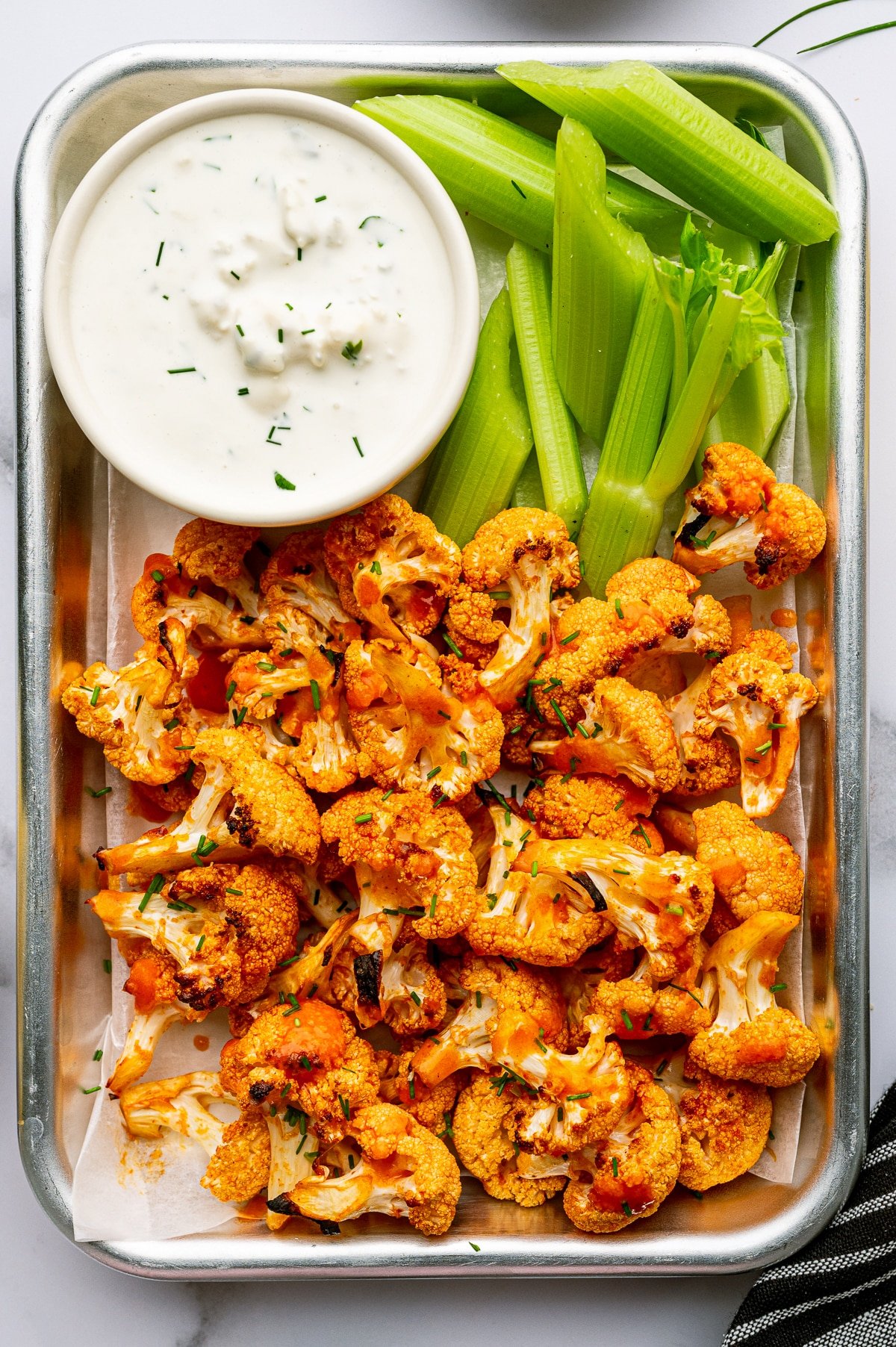 Overhead serving photo of baked buffalo cauliflower in a small metal tray lined with parchment paper, topped with chopped green chives.