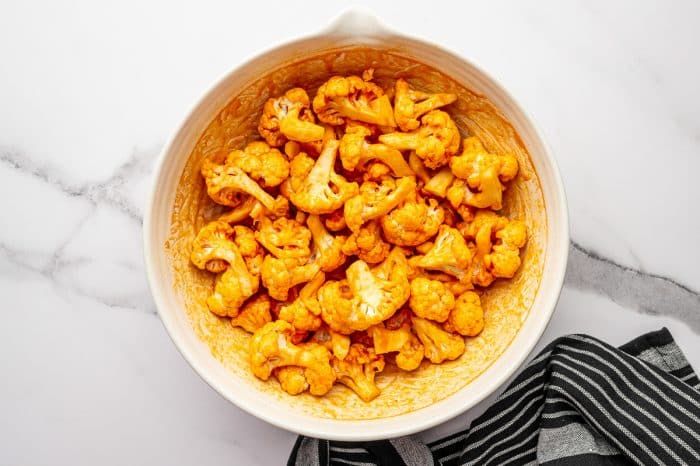 Overhead photo of cauliflower florets fully coated in bright orange buffalo sauce inside a large light beige mixing bowl, with sauce streaks around the sides of the bowl and a black-and-white striped towel to the right.