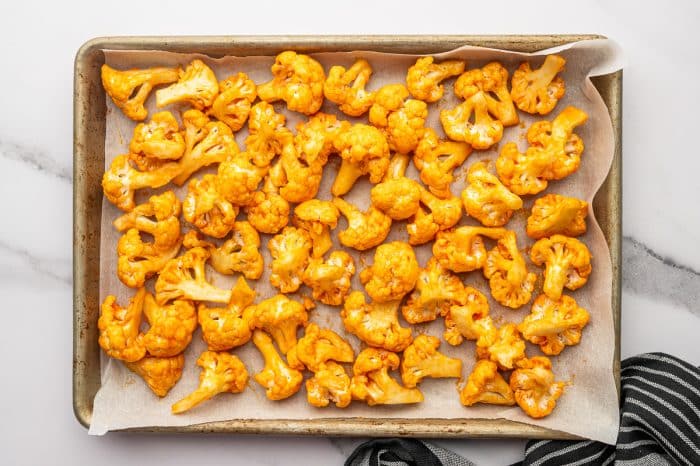 Overhead photo of buffalo-coated cauliflower florets spread in a single layer on a parchment-lined metal baking sheet before baking, with a black-and-white striped towel in the bottom right corner.