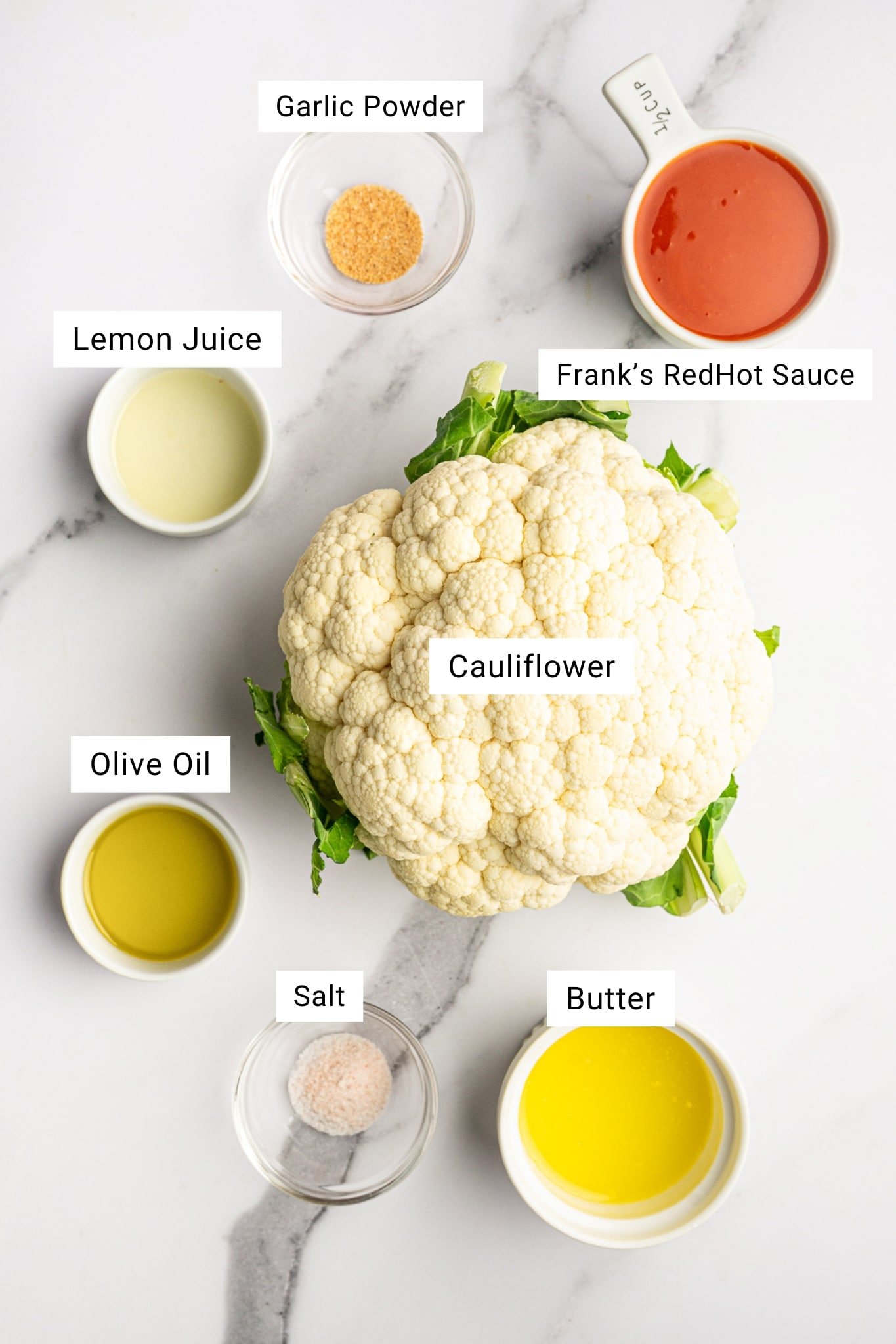 Overhead ingredients photo for baked buffalo cauliflower on a white marble surface with a whole head of cauliflower, a white measuring cup of Frank’s RedHot sauce labeled “Frank’s RedHot Sauce,” a small bowl of melted butter labeled “Butter,” a small bowl of olive oil labeled “Olive Oil,” a small bowl of lemon juice labeled “Lemon Juice,” a small bowl of garlic powder labeled “Garlic Powder,” and a small glass bowl of salt labeled “Salt,” with white text overlay labels in black font.