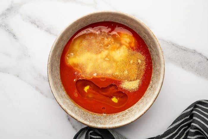 Overhead photo of buffalo sauce ingredients in a tan ceramic bowl on a white marble surface, showing Frank’s RedHot sauce with melted butter pooling on top, olive oil, and garlic powder sprinkled over the surface, with a black-and-white striped kitchen towel in the bottom right corner.