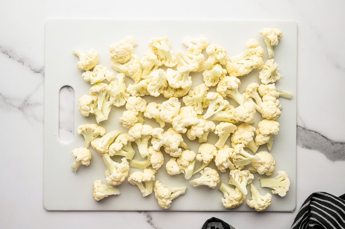 Overhead photo of raw cauliflower florets spread across a large white cutting board on a light marble countertop, with a black-and-white striped kitchen towel in the bottom right corner.