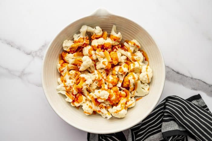Overhead photo of raw cauliflower florets in a large light beige mixing bowl with buffalo sauce drizzled over the top, set on a white marble surface with a black-and-white striped towel on the right side.
