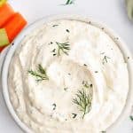 White bowl filled with creamy dill dip served with carrot and celery sticks and salty chips.