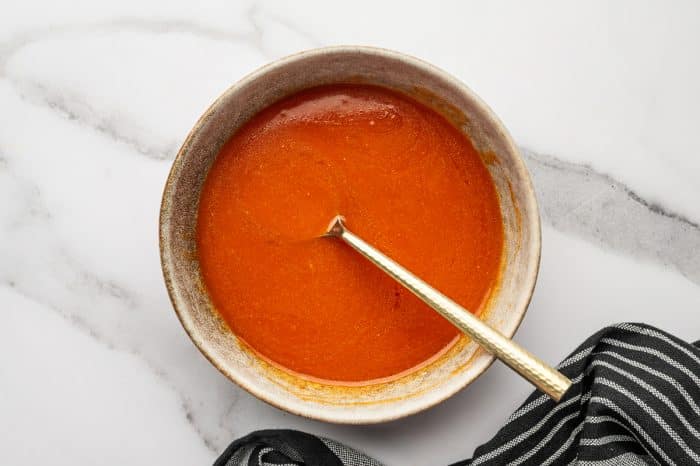 Overhead photo of smooth homemade buffalo sauce in a tan ceramic bowl with a gold spoon resting inside, set on a white marble surface with a black-and-white striped towel in the bottom right corner.
