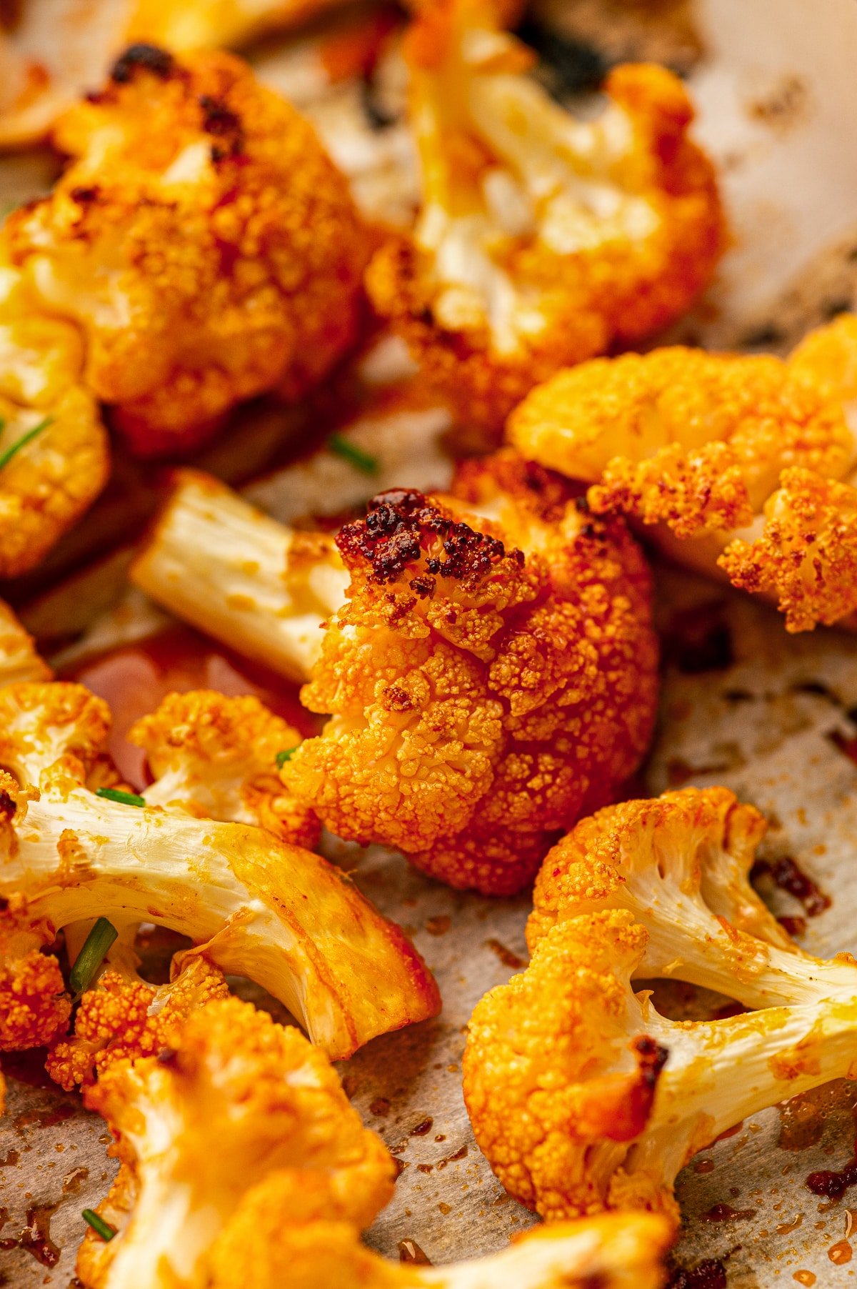 Close-up photo of baked buffalo cauliflower florets on parchment paper, showing bright orange buffalo sauce coating with browned, slightly charred spots on the cauliflower tops.
