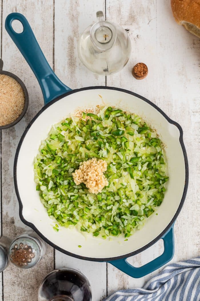 Minced garlic added to a large skillet filled with finely diced onion and green bell pepper cooking in butter.