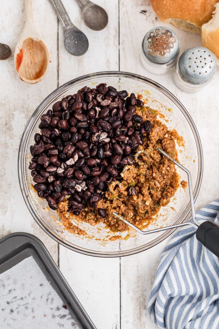 Dried out black beans added to a large glass mixing bowl filled with mashed mixture of softened onion and bell pepper, minced garlic, garlic powder, smoked paprika, Chipotle chili powder, cumin, breadcrumbs, eggs, ketchup, Worcestershire Sauce, salt and pepper.