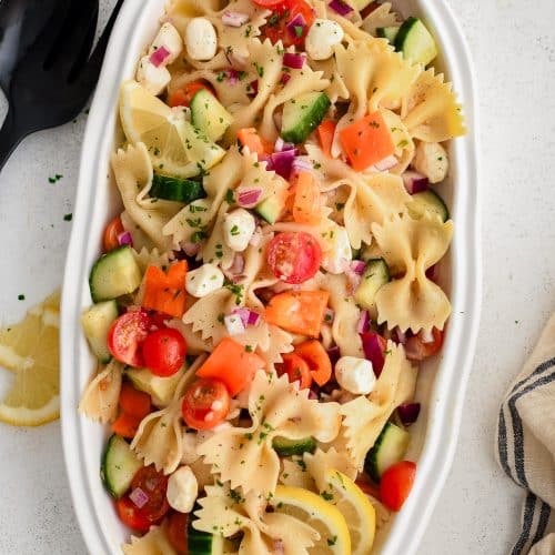 Large white oval-shaped serving plate filled with bowtie pasta, cherry tomatoes, cucumber, diced red onion, and mozzarella pearls all tossed in a simple champagne vinaigrette.