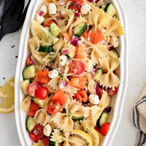 Large white oval-shaped serving plate filled with bowtie pasta, cherry tomatoes, cucumber, diced red onion, and mozzarella pearls all tossed in a simple champagne vinaigrette.