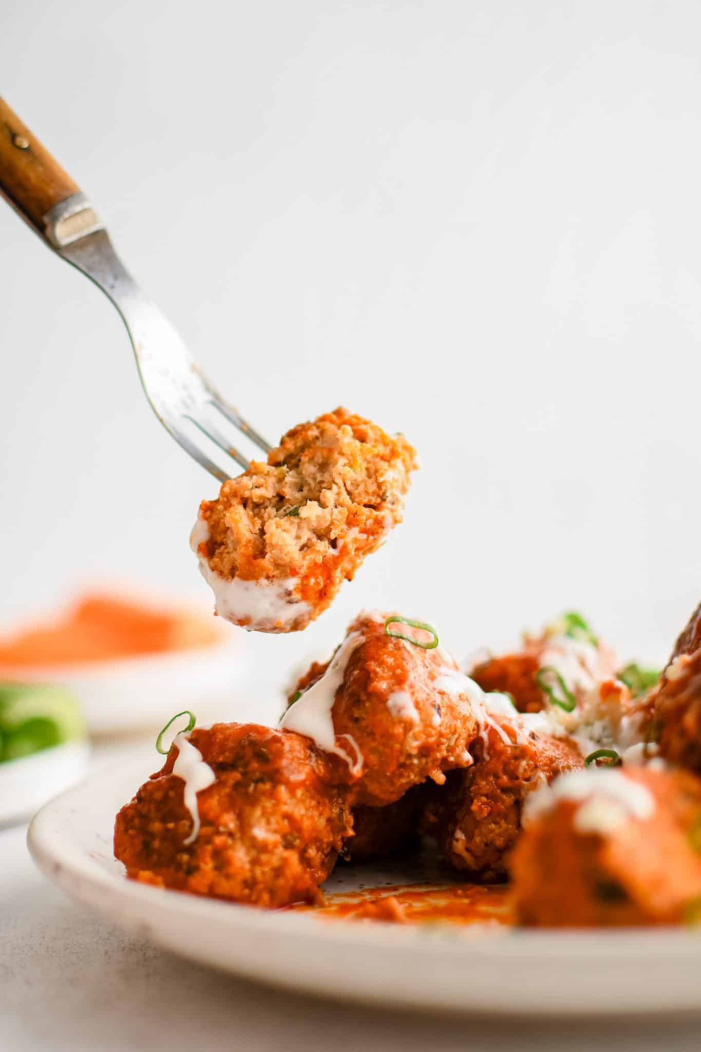 Fork with half of a buffalo chicken meatball hovering above a large plate filled with the remaining meatballs.