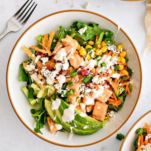 Assembled buffalo chicken chopped salad made with Romaine lettuce topped with creamy buffalo sauce coated chicken, avocado, corn, carrots, celery, green onions, blue cheese, tortilla strips, cooked bacon, and drizzled with creamy ranch dressing.