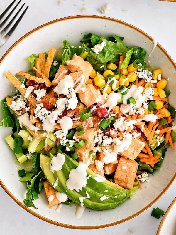 Assembled buffalo chicken chopped salad made with Romaine lettuce topped with creamy buffalo sauce coated chicken, avocado, corn, carrots, celery, green onions, blue cheese, tortilla strips, cooked bacon, and drizzled with creamy ranch dressing.