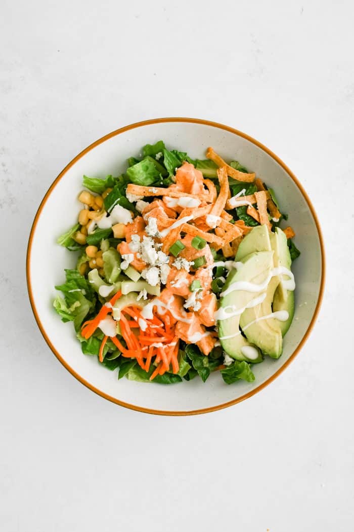 Large white salad bowl with chopped Romaine lettuce as a base topped with chicken, avocado, corn, carrots, celery, green onions, blue cheese, tortilla strips, cooked bacon, and ranch dressing.