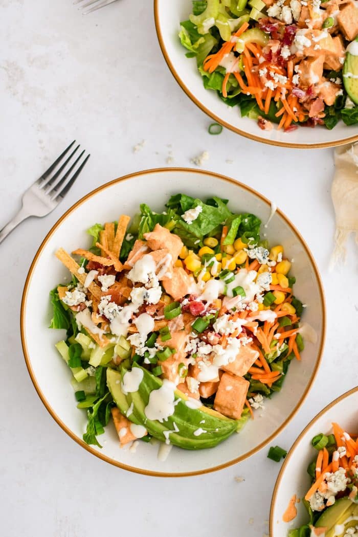 Assembled buffalo chicken chopped salad made with Romaine lettuce topped with creamy buffalo sauce coated chicken, avocado, corn, carrots, celery, green onions, blue cheese, tortilla strips, cooked bacon, and drizzled with creamy ranch dressing.