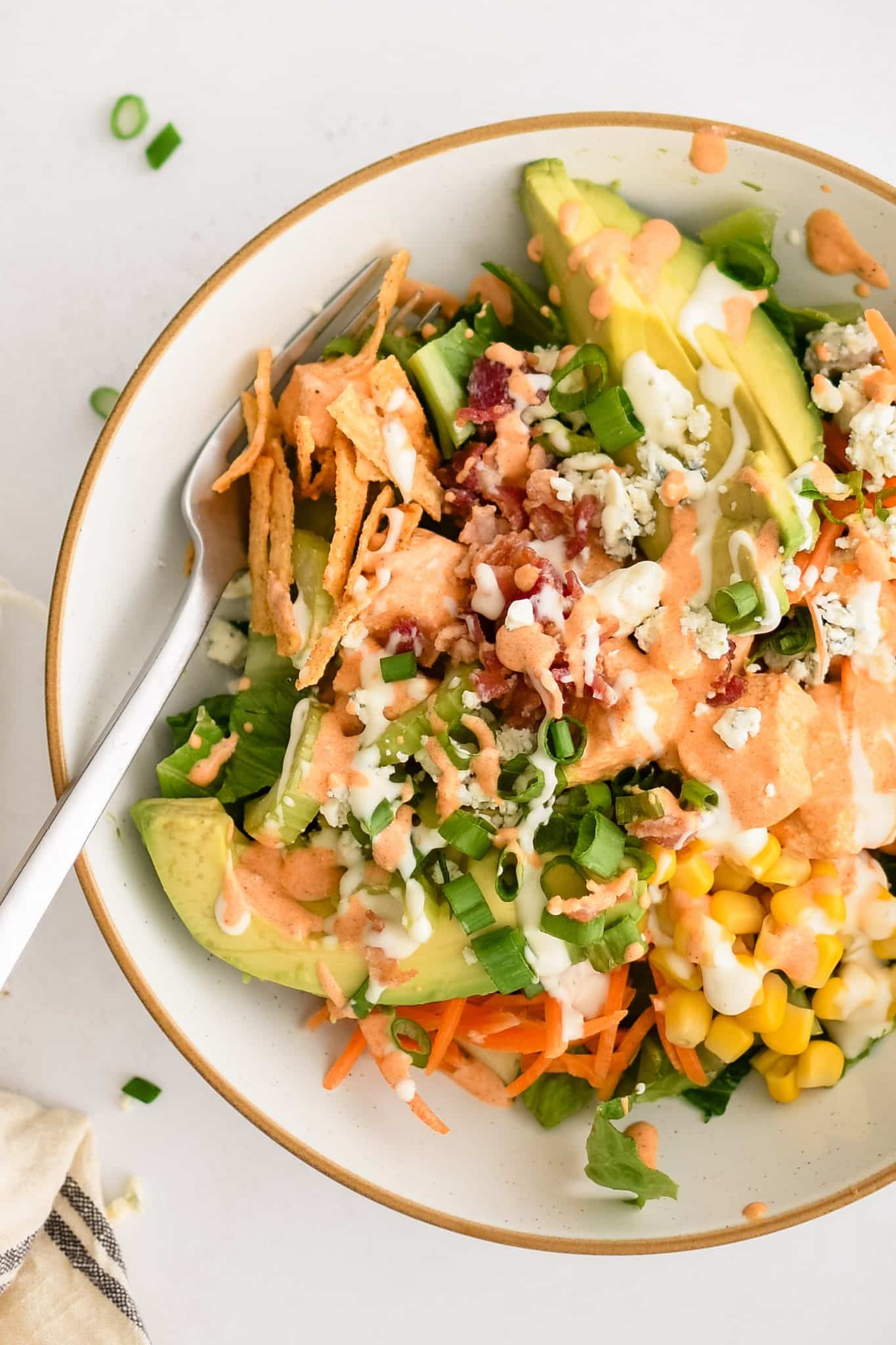 Assembled buffalo chicken chopped salad made with Romaine lettuce topped with creamy buffalo sauce coated chicken, avocado, corn, carrots, celery, green onions, blue cheese, tortilla strips, cooked bacon, and drizzled with creamy ranch dressing.