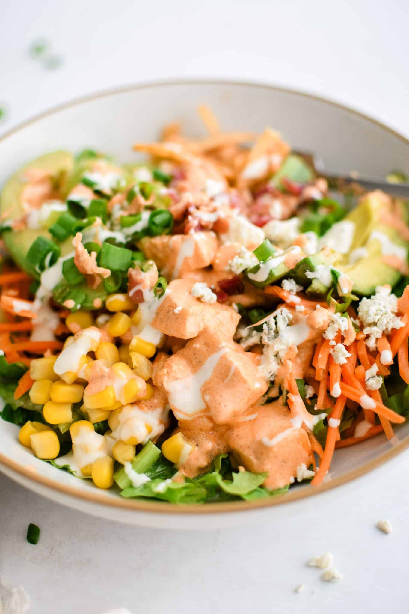 Assembled buffalo chicken chopped salad made with Romaine lettuce topped with creamy buffalo sauce coated chicken, avocado, corn, carrots, celery, green onions, blue cheese, tortilla strips, cooked bacon, and drizzled with creamy ranch dressing.