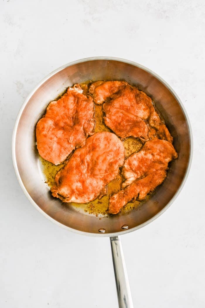 Four thinly sliced chicken breasts covered in seasoning cooking in a large stainless steel skillet.