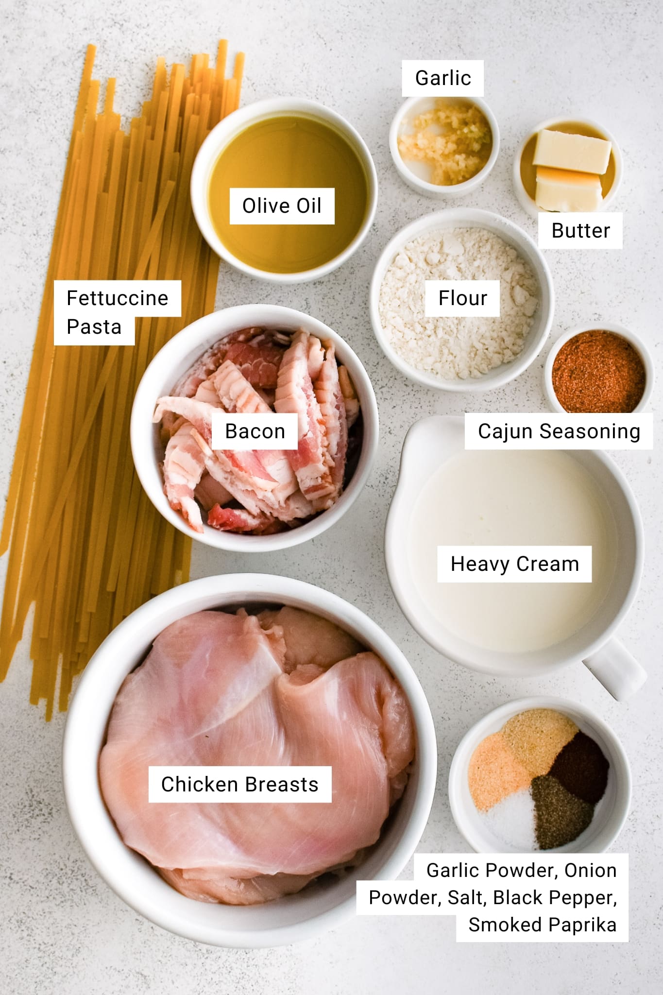 Overhead image of labeled ingredients for Cajun Chicken Alfredo, including fettuccine pasta, chicken breasts, bacon, olive oil, butter, garlic, flour, Cajun seasoning, heavy cream, and a spice blend of garlic powder, onion powder, salt, pepper, and smoked paprika.