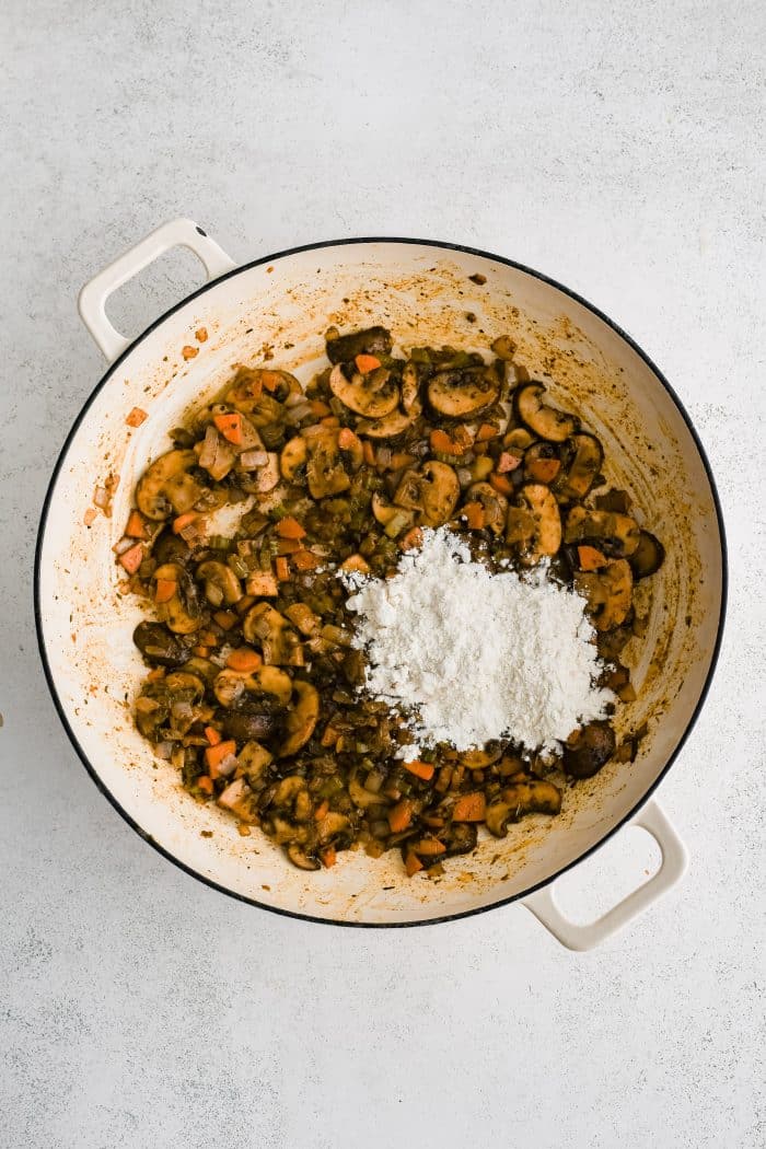 Flour sprinkled over browned and softened seasoned mushrooms, onions, celery, and carrots, sauteing in a large white oven-safe pan.