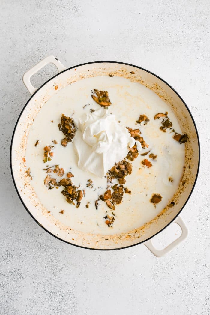 Chicken broth, heavy cream, and sour cream added to the combination of sauteed onions, carrots, celery, and mushrooms coated in seasoning and all-purpose flour cooking in a large skillet.