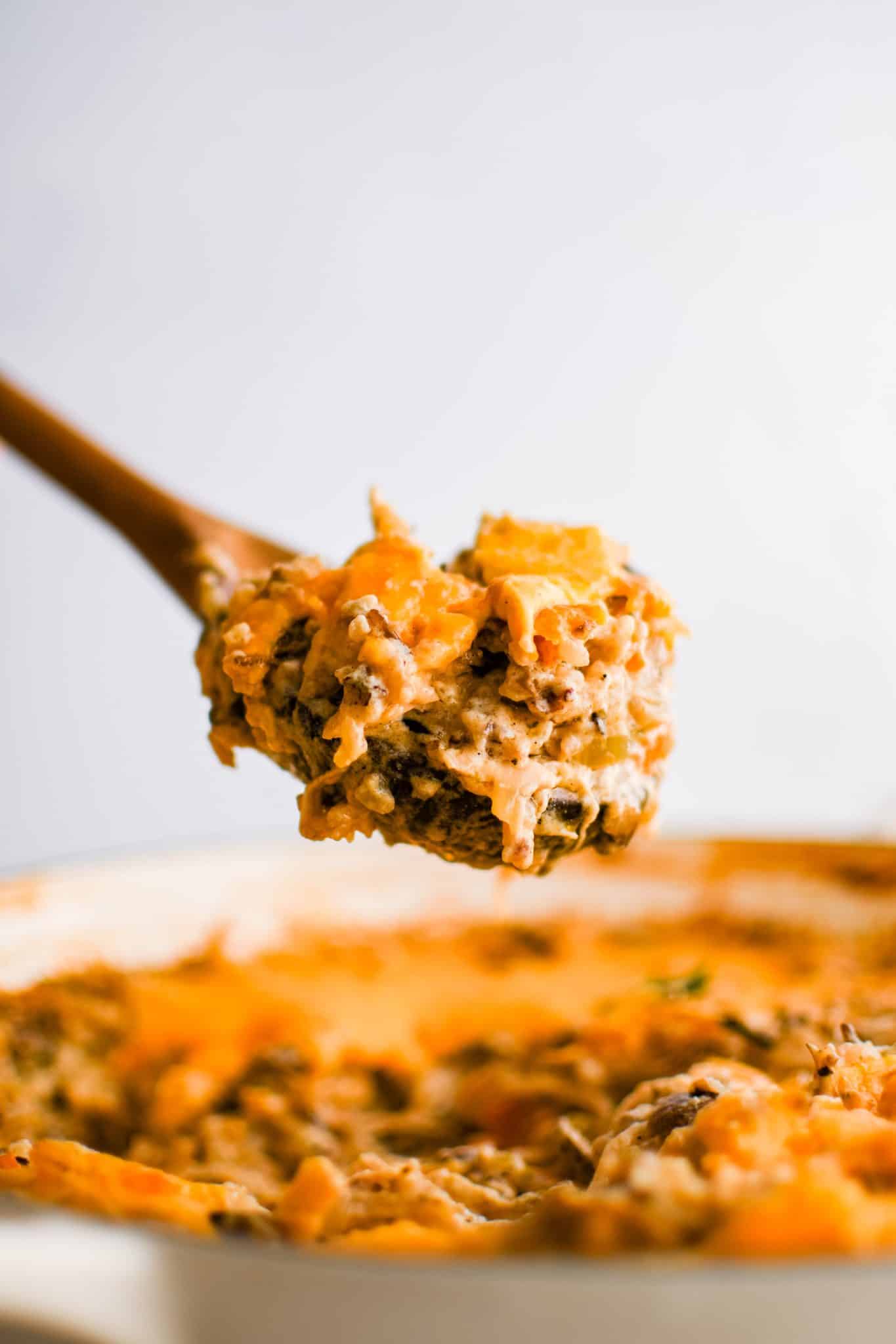 Large wooden spoonful of chicken and wild rice casserole hovering over a large pan filled with cheesy chicken and rice.