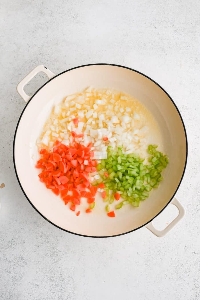 Large white oven-safe skillet filled with chopped onions, carrots, and celery cooking in butter.