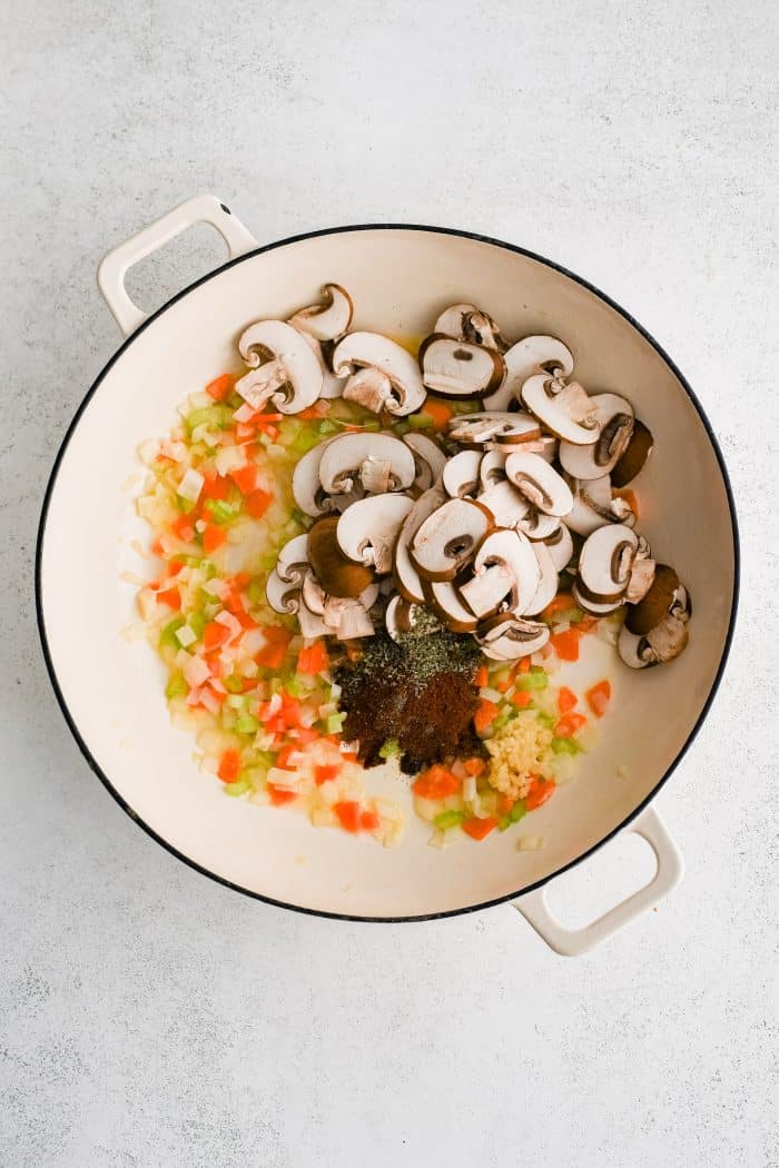 Mushrooms, garlic, paprika, Italian seasoning, salt, and pepper added to a large white oven-safe skillet filled with sauteing chopped onions, carrots, and celery.