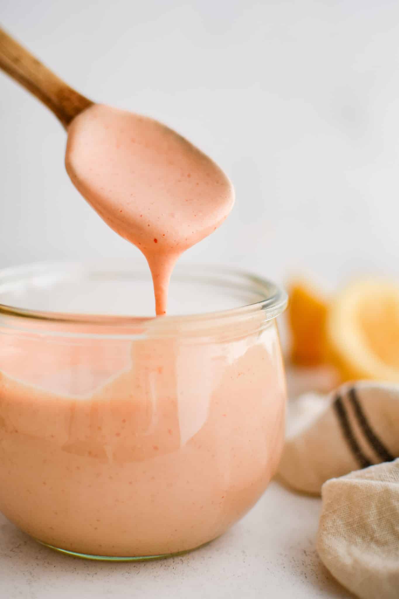 Homemade Mississippi Comeback Sauce dripping off a small wooden spoon into a small glass jar filled with the remaining comeback sauce.