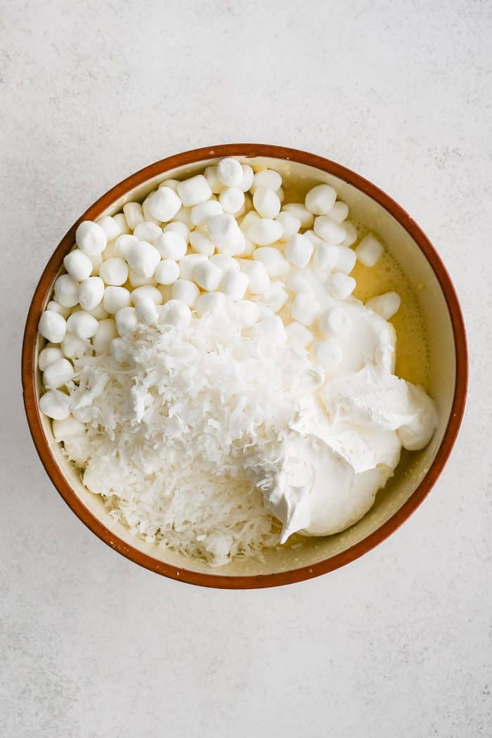 Whipped topping, marshmallows, and coconut added to a large mixing bowl filled with whipped together cream cheese, instant vanilla pudding mix, and crushed pineapple.