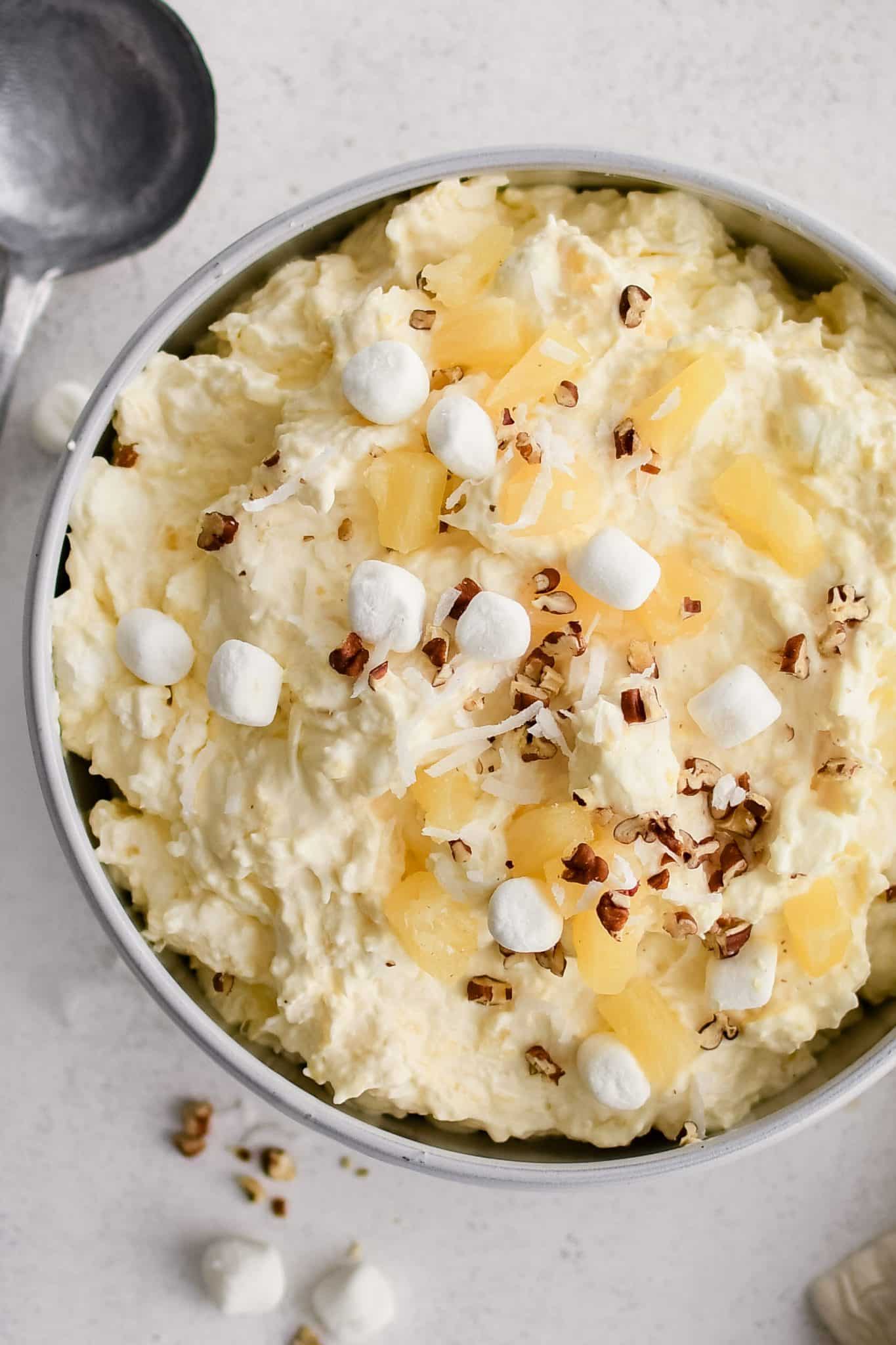 Large white serving bowl filled with prepared creamy pineapple fluff dessert topped with pineapple chunks, mini marshmallows, and chopped pecans.