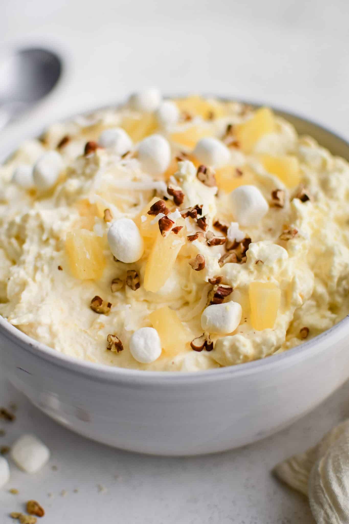 Large white serving bowl filled with prepared creamy pineapple fluff dessert topped with pineapple chunks, mini marshmallows, and chopped pecans.