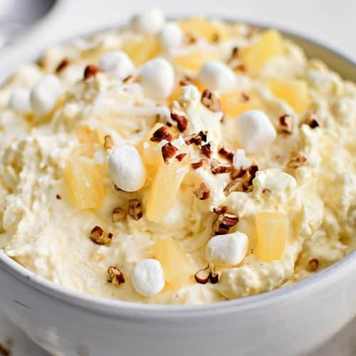 Large white serving bowl filled with prepared creamy pineapple fluff dessert topped with pineapple chunks, mini marshmallows, and chopped pecans.