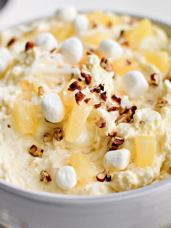 Large white serving bowl filled with prepared creamy pineapple fluff dessert topped with pineapple chunks, mini marshmallows, and chopped pecans.