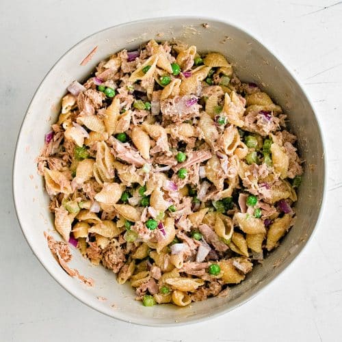 Large white mixing bowl filled with prepared tuna pasta salad made with cooked shells, peas, diced celery and onion, in a creamy mayonnaise-based dressing.