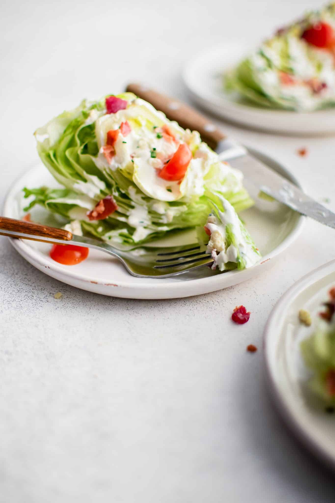 Serving plates each with one quarter of an iceberg lettuce topped with homemade blue cheese dressing and sprinkled with crumbled bacon bits, halved cherry tomatoes, chives, and blue cheese crumbles.