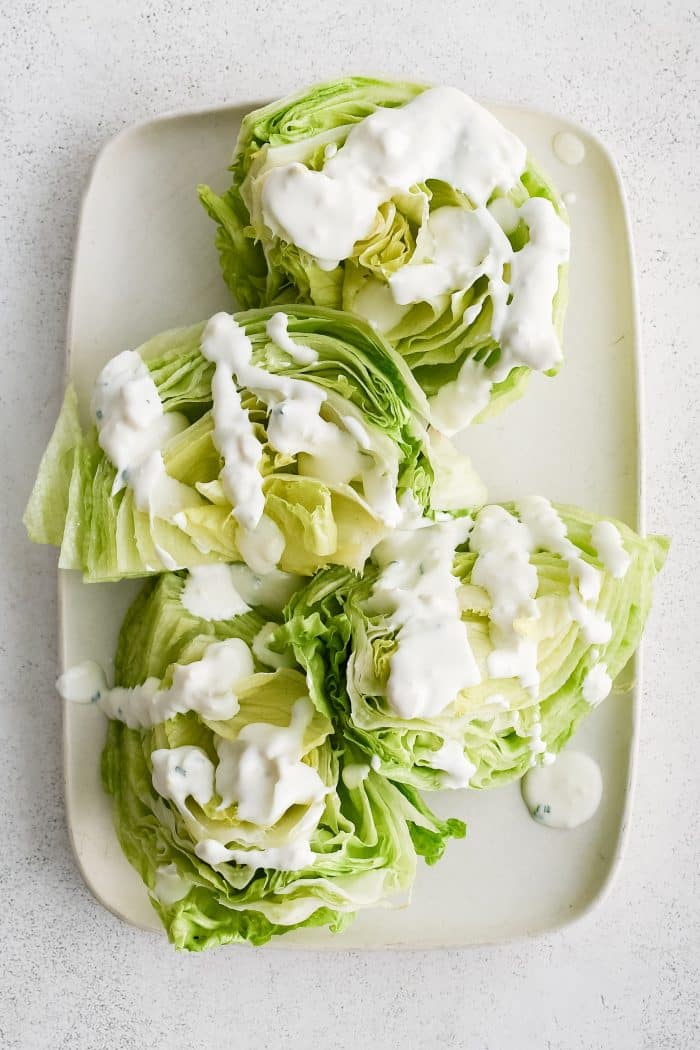 Large white rectangular serving platter with four wedges of iceberg lettuce, cut-side-up, drizzled with homemade blue cheese dressing.